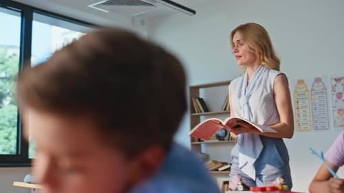 School Teacher Walking Classroom with Book Observing Studying Pupils Writing