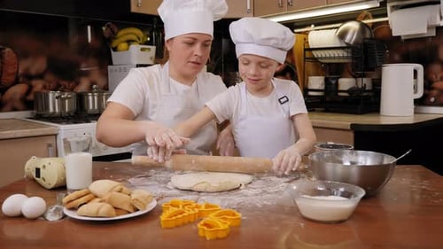 A Boy in an Apron and a Cap Rolls Out the Dough with a Rolling Pin with Mother