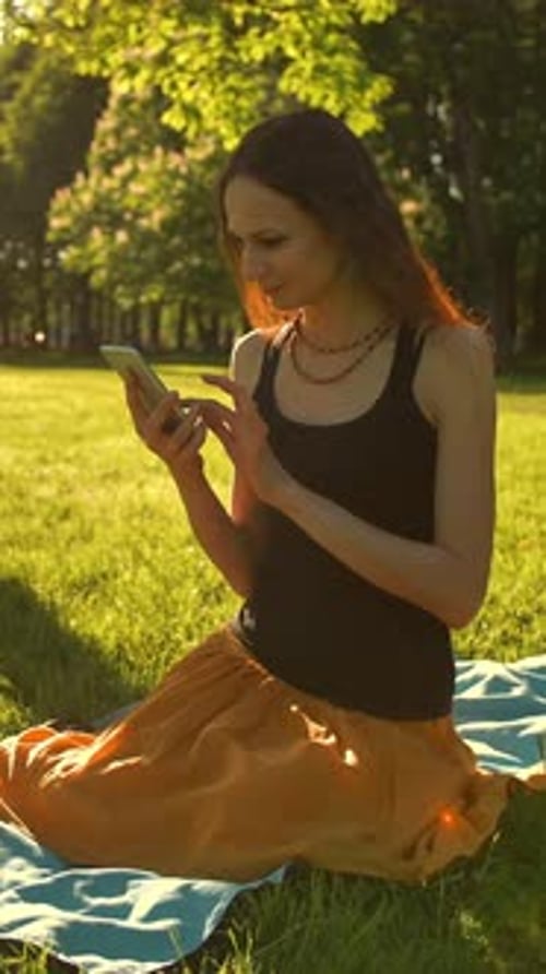 Vertical of Young Lady Enjoying Nature and Sunset Sitting on Green Summer Park Using Mobile