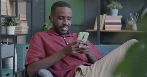 Fröhlicher Student, der Smartphone-SMS in sozialen Medien nutzt, sitzt im Sessel in der Wohnung