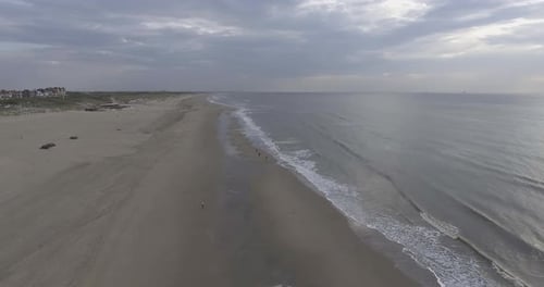 A drone shot by the sea, panning right, waves rolling in and people walking on the beach