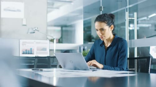 Beautiful Female Data Analyst sitting at the Table Works on a Laptop. Stylish Woman in the Modern O