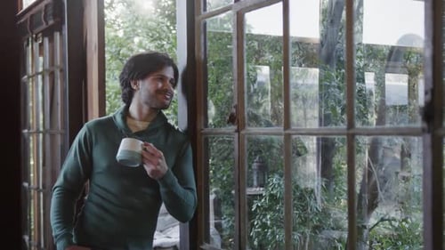 Happy caucasian man with coffee and looking out window at home, copy space, slow motion