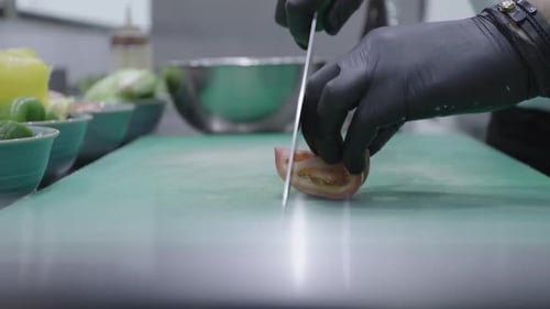 Chef cutting a red tomato using a knife with black gloves on a green cutting board.