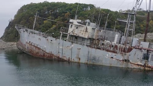 Rostgraues, halb untergetauchtes Schiffswrack, das an einem bewölkten, bewölkten Tag in Küstennähe vor Anker lag. Langsames Antennenkabel