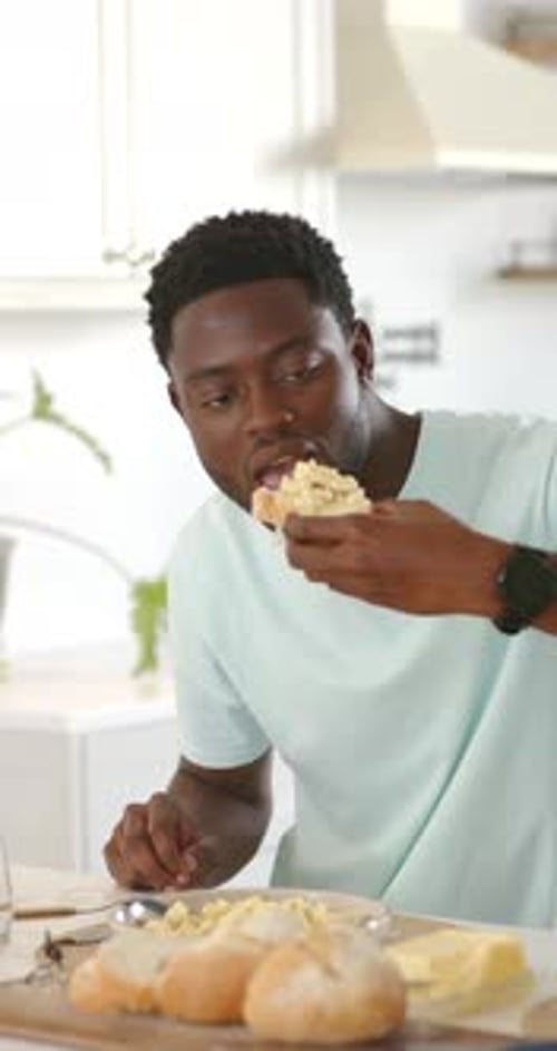 Vidéo verticale : un homme afro-américain ramasse un petit pain dans une assiette dans la cuisine, mordant de la pâte à tartiner