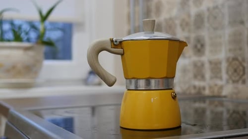 A Yellow Coffee Maker Sits on a Stove with a Plant and Window in the Background When It Boils It is