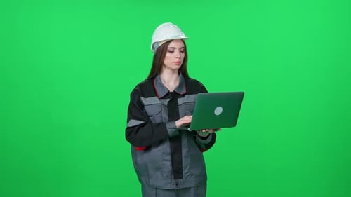 Focused Woman Worker in a Uniform and Protective Helmet Technical Engineer Stands and Uses a Laptop