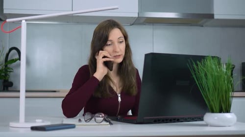 Using Laptop and Sitting at Home Office Desk Young Businesswoman Talks on Phone with Smile As She