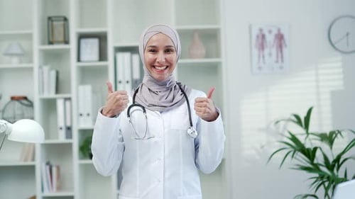 Happy muslim female doctor showing thumbs up gesture while standing in office in modern hospital cli