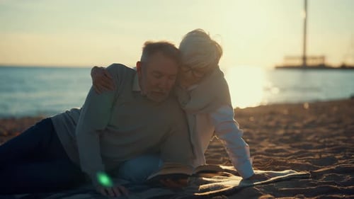 Senior Gray Haired Man Woman Reading Book Together Sitting on Sandy Sea Beach at Sunset in Evening