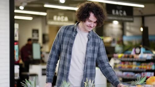 Happy Brunette Guy with Curly Hair in a Plaid Shirt Examines Products in the Citrus Department of