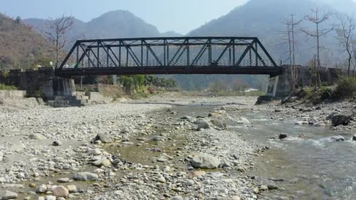 Aerial drone footage of a steel truss bridge spanning a rocky riverbed surrounded by scenic