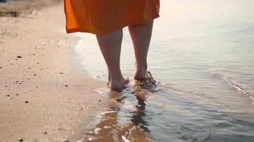 Legs of Woman in Orange Dress Walking on Sand on Beach Washed By Sea Waves