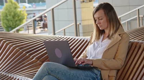 Woman Sitting on Bench in City Street and Using Laptop