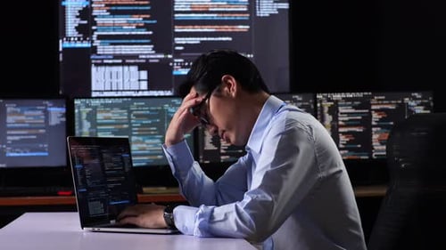 Vista lateral del programador que tiene dolor de cabeza mientras escribe código con una computadora portátil usando varios monitores