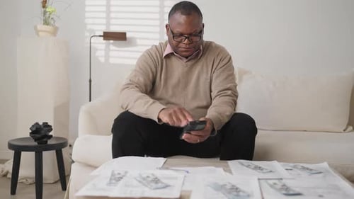 Man Sitting on Couch Reviewing Documents and Using Phone