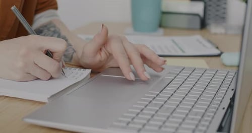 Closeup of Hand with Tattoo Using Laptop and Writing Information in Notebook Working at Desk