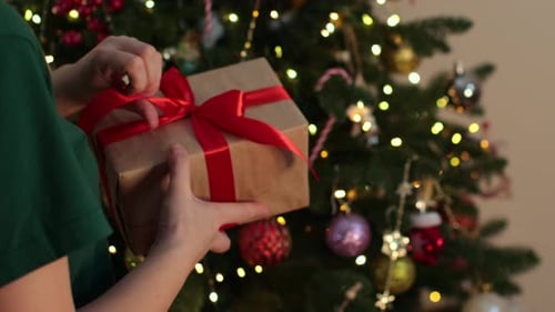 Person Unties Ribbon on Christmas Gift by Tree