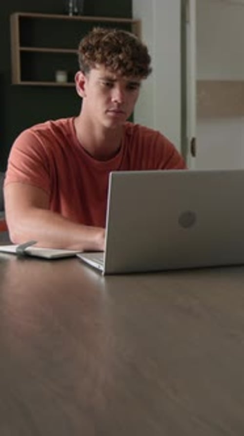 Vertical video: Entering home office, man in twenties studying at desk with laptop writing notes