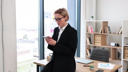 Businesswoman Browsing on Phone While Leaning on Office Desk During Work