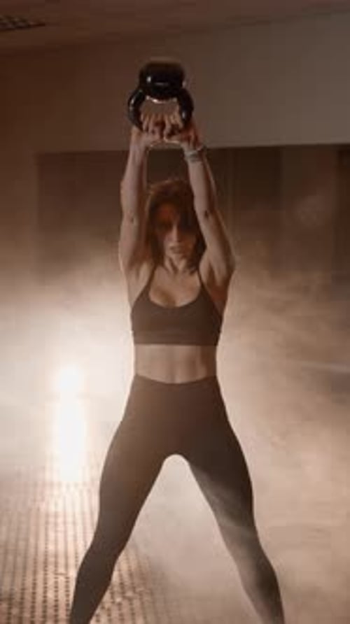 Athletic Woman in Sportswear Performing Kettlebell Swing Exercise in a Smoky Gym Environment Focused
