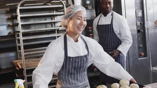 Happy diverse bakers working in bakery kitchen, holding baking sheet with rolls in slow motion
