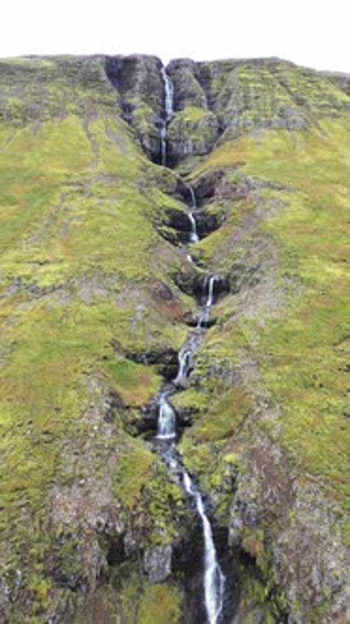 Aerial drone view of a long cascading waterfall flowing down a steep green mountainside in Iceland.