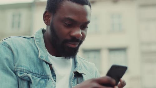 Close Up Happy Young African American Man Uses the Phone Sitting Outdoor at the Street Business