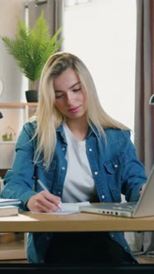 Woman Taking Notes at Table While Using Laptop