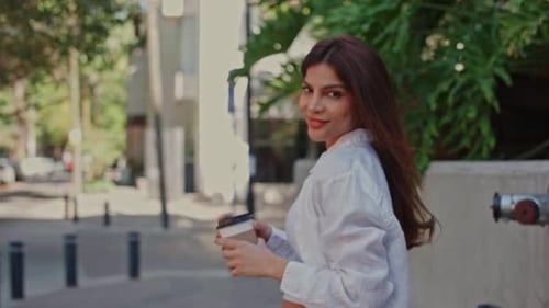 Woman Walking on City Street and Sipping Coffee While Looking at Camera