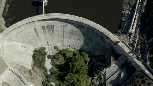 Toma aérea mirando directamente hacia abajo, volando sobre una gran presa curva de hormigón, se desliza sobre un rojo oscuro y vítreo