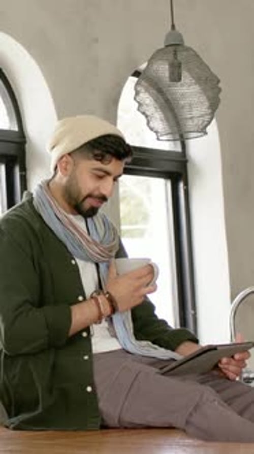 Vertical video: Glancing at tablet, man in beanie and scarf sipping white mug to read at wood bar