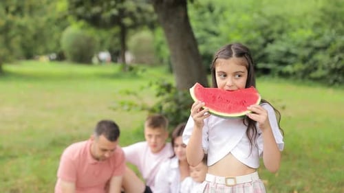 A Little Girl Eats a Slice of Watermelon in a Summer Park Family in the Background