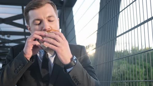 Young Successful Businessman in Black Suit Walking Near Office Building and Eating Fast Food