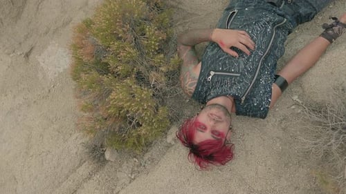 A man with red hair and red eye makeup lies on a rocky desert looking upward