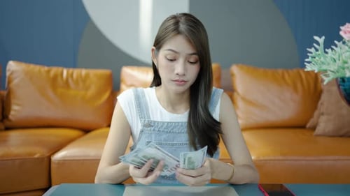 Upset young asian woman counting money on sofa, Sad girl model holding money cash in home.