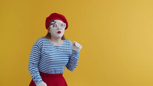 Eine junge Pantomime in leuchtenden Kleidern legt ihre Hand ans Ohr und tut so, als würde sie Klatsch und Tratsch belauschen und