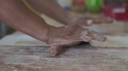 Close-up of a pastry chef rolling dough with hands using a wooden rolling pin on a work surface.