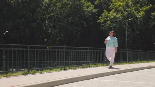 A beautiful woman in a striped shirt and white pants walks alone. A woman holds a phone in her hands