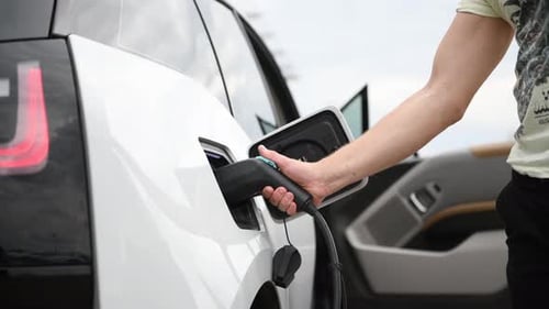 Woman stands with phone near her electric car and waits when vehicle will charged, then closes trunk