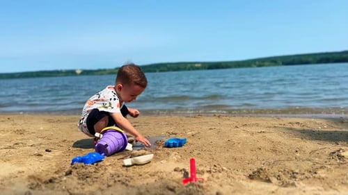 Serious baby boy sits squatted on the beach. Toddler plays with toy in the sand near the river.