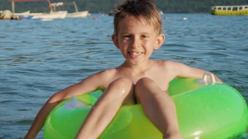 Happy smiling boy sitting in green inflatable ring and swimming in the sea towards camera