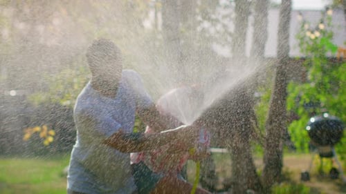 Happy Young Couple Has Fun on a Hot Summer Day Playing with Water Hose Sprinkler in the Garden. Wat