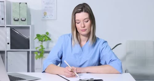 Doctor in scrubs with stethoscope working with laptop and clipboard at white table in clinic
