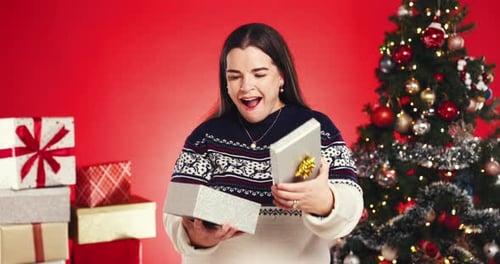 Happy woman, face and Christmas surprise with gift box in studio for giving on a red background