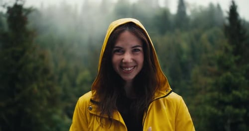 A Happy Girl in a Yellow Jacket with a Hood Stands Against the Backdrop of a Coniferous Mountain