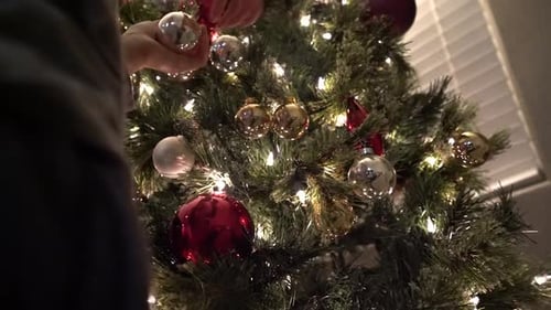 Decorating Christmas tree. Close-up. Boy placing festive ornaments on a lit up Christmas tree. 4K.
