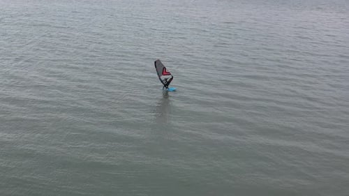 An aerial view of a windsurfer gliding across the water's surface. The windsurfer is wearing a wetsu