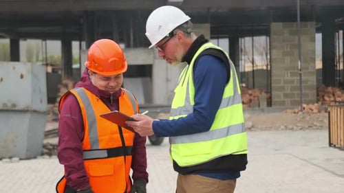 Handwerker mit orangefarbenem Helm, Weste und Schutzhandschuhen kommuniziert mit dem Bauingenieur